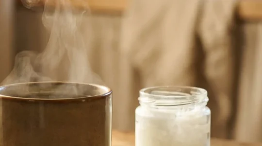 Tasse fumante d'infusion au CBD posée près d'une cuillère d'huile de coco et de fleurs de chanvre séchées, dans une lumière dorée naturelle